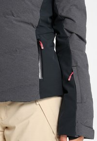 Gros plan d'une personne portant une veste grise et noire avec des tirettes de fermeture éclair roses et un pantalon beige, montrant la poche zippée et les détails des manches.