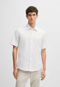 Camicia bianca a maniche corte con colletto appuntito, realizzata in tessuto leggero, con un logo discreto e un orlo curvo.