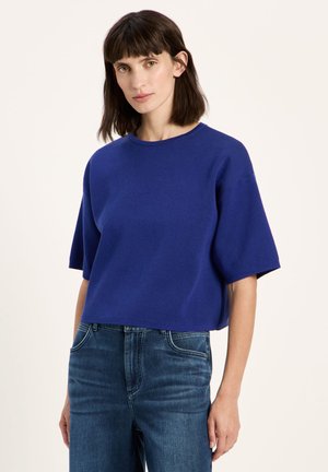 Woman with straight dark brown hair wears a loose short-sleeve cobalt blue top and high-waisted blue jeans against a plain background.