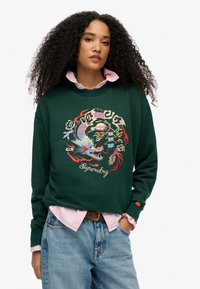 Groene sweatshirt met ingewikkelde rode en blauwe draakbrodering, met "Superdry" tekst, gelaagd over een lichtroze overhemd met kraag.