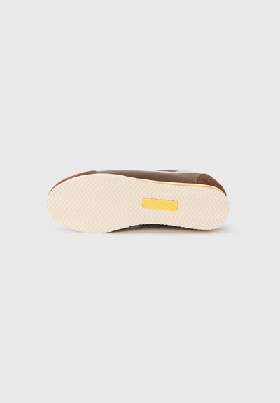 Adidași maro cu o parte superioară texturată, talpă din cauciuc bej și etichetă galbenă accent, având un model de rulare zigzag pe partea de jos.