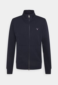 Giacca a zip blu navy realizzata in tessuto morbido, con colletto alto, tasche frontali e un piccolo logo sul petto. Polsini e orlo a coste.