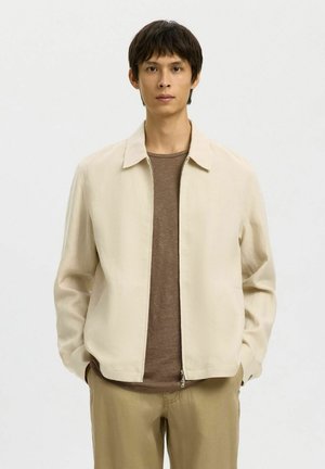 Junger Mann trägt eine beige Jacke mit Reißverschluss über einem braunen Hemd und beigen Hosen und steht mit den Händen in den Taschen vor einem einfarbigen Hintergrund.