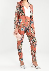 Květinový kostým v korálové barvě s modrými a žlutými květy; saka na míru s dvojitými knoflíky, zúžené kalhoty, dopln�ěné stříbrnými mokasínami a růžovou kabelkou.