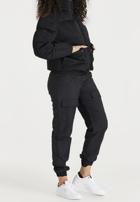 Svart puffjacka med hög krage och fickor fram, tillsammans med svarta cargobyxor med fickor och elastiska muddar, samt vita sneakers.