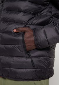 Giacca a vento nera con design trapuntato, dotata di tasca laterale con cerniera, materiale leggero e polsini elasticizzati per una vestibilità aderente.