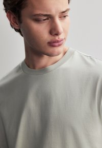 Hellgraues T-Shirt mit kurzen Ärmeln und rundem Ausschnitt, hergestellt aus weichem Stoff, mit glatter Textur und minimalistischem Design.