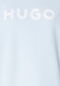 Camiseta de color azul claro hecha de tela suave con un gran bordado en blanco que dice "HUGO". Diseño simple con una textura suave.