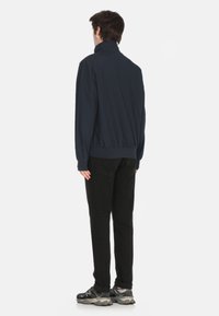 Navy bomber jacket with high collar, ribbed hem and cuffs, paired with black pants and gray sneakers, viewed from the back.