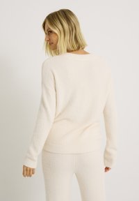 Pull beige clair duveteux avec un col rond, des poignets et un ourlet côtelés. Le tissu semble doux et texturé, conçu pour une coupe décontractée.
