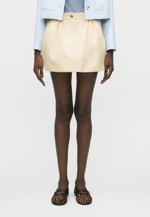 Jambes portant des collants foncés, une mini-jupe beige avec des poches, une veste courte bleu clair et des ballerines Mary Jane à motif léopard.