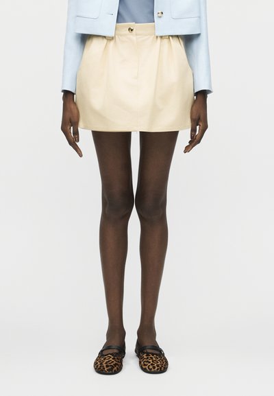 Jambes portant des collants foncés, une mini-jupe beige avec des poches, une veste courte bleu clair et des ballerines Mary Jane à motif léopard.
