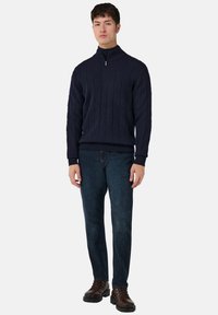 Mladý muž stojící v námořnickém svetr s polovičním zipem a pleteným vzorem, tmavých džínách a hnědých kožených botách na jednoduchém světlém pozadí.