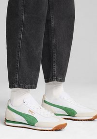 Zapatillas blancas y verdes con materiales de ante y malla. Presentan una suela de goma y una marca destacada en el costado. Combinadas con pantalones de mezclilla oscura.