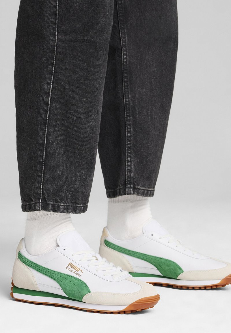 Zapatillas blancas y verdes con materiales de ante y malla. Presentan una suela de goma y una marca destacada en el costado. Combinadas con pantalones de mezclilla oscura.