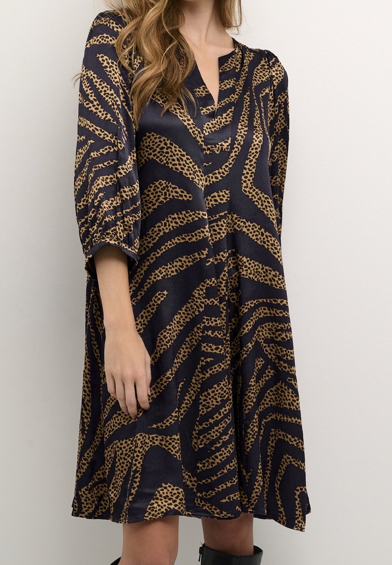 Marindräkt i marinblått med tan leopardmönster i ett silkesliknande material. Har V-ringning, ¾ ärmar och en lös A-linjeform. Minimalistisk design.