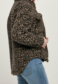 Osoba w brązowo-czarnym futrzanym kurtce w wzór lamparta, nosząca niebieskie dżinsy, przedstawiona z boku na jednolitym tle.