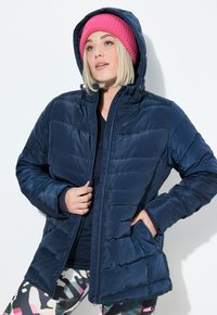 Chaqueta de plumas navy con cierre de cremallera y capucha. Presenta secciones acolchadas y bolsillos laterales. Lleva puesta sobre leggings estampados y una camiseta oscura.