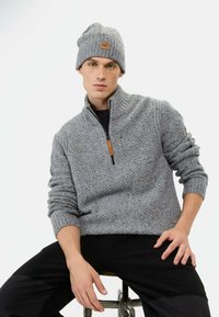 Grijze gebreide sweater met een hoge kraag en halve rits, gecombineerd met een bijpassende geribbelde beanie. Bevat een leren accent op de sweater.