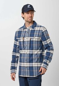 Homme portant une chemise à carreaux bleue et blanche à boutons, un pantalon foncé avec des poches zippées et une casquette noire avec un logo élan blanc.