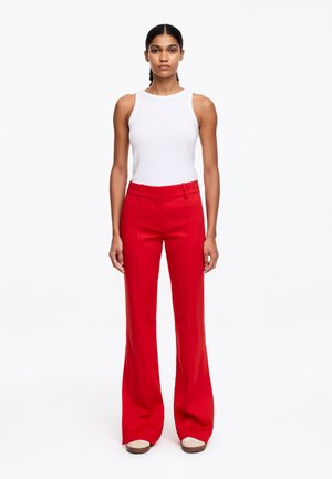 Woman standing wearing a white sleeveless top and high-waisted red wide-leg trousers with beige shoes, against a plain white background.