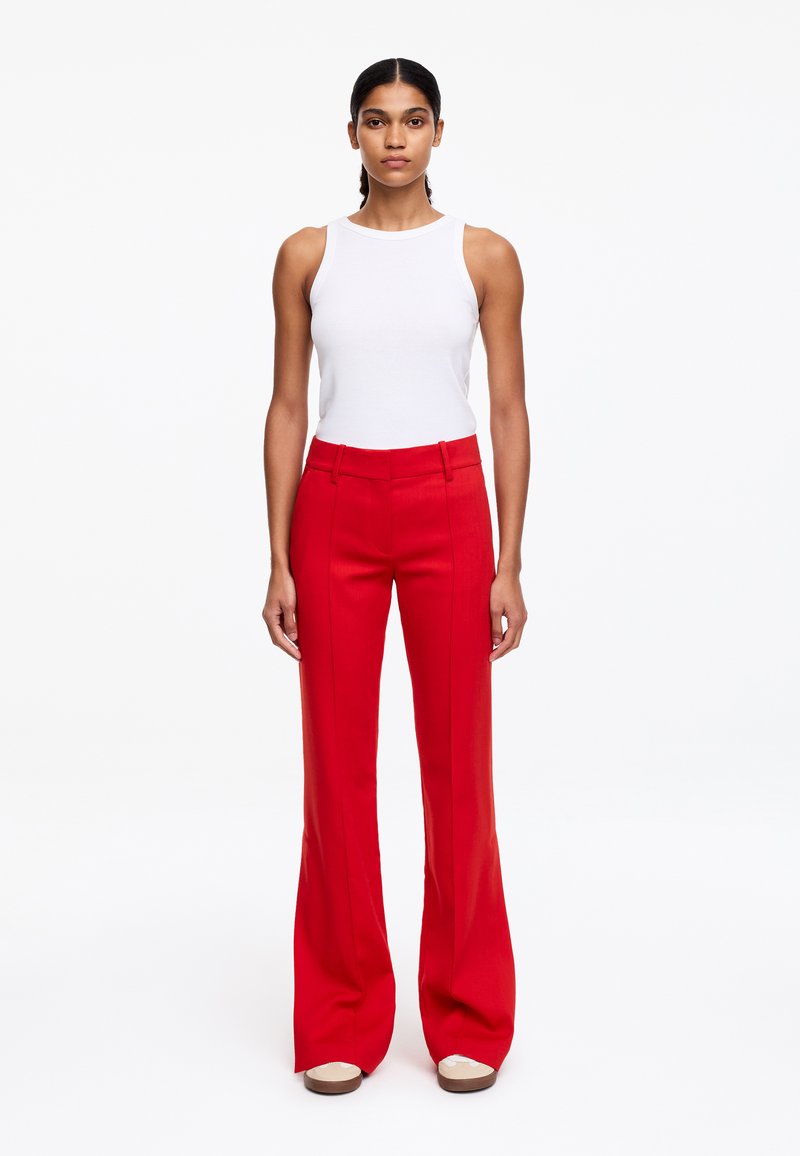 Femme debout portant un haut blanc sans manches et un pantalon large taille haute rouge avec des chaussures beige, sur un fond blanc uni.