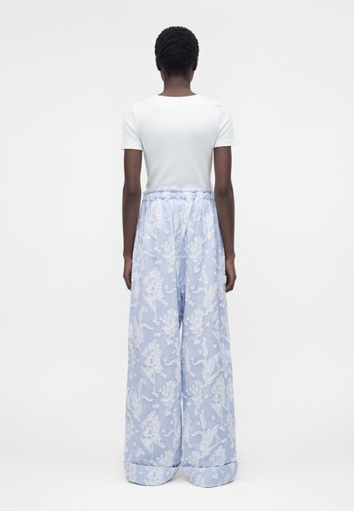 Άτομο με κοντά σγουρά μαλλιά που φοράει εφαρμοστό λευκό κοντομάνικο τοπ και φαρδιά ανοιχτομπλε παντελόνια με λευκά λουλουδάτα σχέδια, στέκεται με την πλάτη προς τα εμπρός.