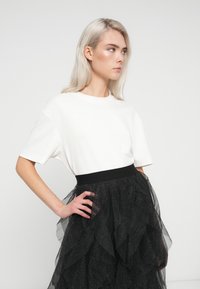 Balta trumpa rankovėmis marškinėliai su laisvu kirpimu, derinami su juoda tiulio sijonu, turinčiu prasišviečiančias sluoksnius ir plačią juodą juostą.