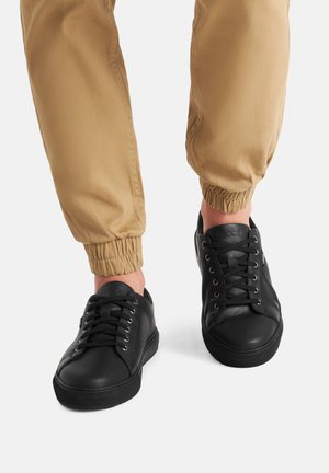 Czarne skórzane sneakersy z okrągłymi noskami, fakturowaną powierzchnią i płaskimi sznurówkami, zestawione z khakimi spodniami z elastycznymi ściągaczami.