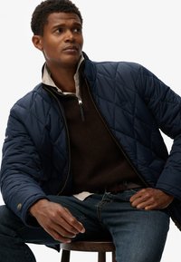 Marineblauwe gewatteerde jas met staande kraag, ritssluiting en ritssluitingaccent. Gelaagd over een bruine gestructureerde trui en een licht shirt. Donkere spijkerbroek gedragen.