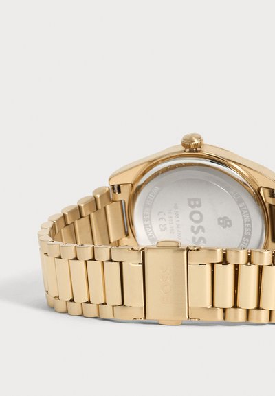Montre-bracelet en métal doré avec fermoir gravé "BOSS" et fond visible marqué.