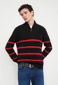 Μαύρο μάλλινο πουλόβερ με κόκκινες οριζόντιες ρίγες, γιακάς με φερμουάρ στο μισό, φορεμένο με μπλε τζιν και μαύρη ζώνη με οπές.