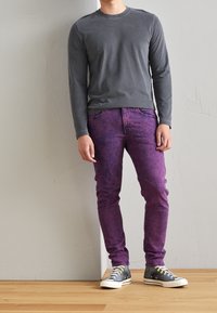 Grå långärmad tröja, lila texturerade jeans och grå sneakers med vita sulor. Fullständig vy av den avslappnade outfiten på en stående figur.