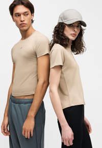Νεαρός άνδρας και γυναίκα στέκονται πλάτη με πλάτη, και οι δύο φορούν μπεζ μπλουζάκια; η γυναίκα φορά ανοιχτό γκρι καπέλο και μαύρο παντελόνι, ο άνδρας φορά γκρι παντελόνι.