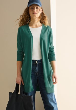 Green, long cardigan with open front, ribbed cuffs, and hem. Worn over a white top, paired with blue denim jeans and a dark tote bag.