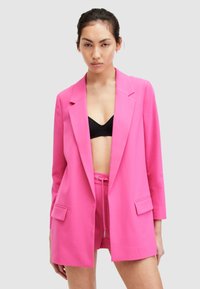 Roza blazer iz lahkega materiala, z izrezom in dvema sprednjima žepoma, v kombinaciji s podloženimi šortkami in črnim braletom.