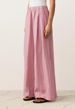 Nő magasderekú, széles szárú rózsaszín lenvászon nadrágot visel gumis derékrésszel és fekete papucs szandállal, világos csempézett padlón állva.