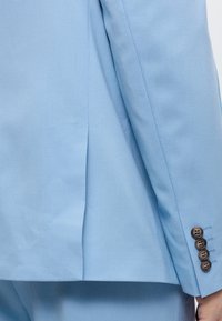 Μανίκι ανοιχτό μπλε σακακιού με τέσσερα σκούρα κουμπιά κοντά στη μανσέτα, που δείχνει μέρος του αντίστοιχου παντελονιού και το χέρι ενός ατόμου.