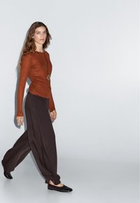 Maglietta a maniche lunghe color ruggine abbinata a un pantalone ampio di un marrone scuro, con una texture liscia. Ballerine nere e una collana con pendente circolare completano l'outfit.