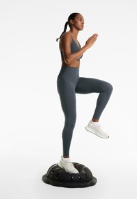 Sötétszürke sportmelltartóval és hozzáillő leggings-szel ellátott sportfelszerelés. Fehér futócipő. Fekete egyensúlyozó tréneren áll.