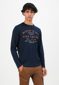 Fiatal férfi göndör hajjal, sötétkék, hosszú ujjú pólót visel narancssárga és fehér felirattal, barna cargo nadrággal párosítva, háttérben fehér fal.