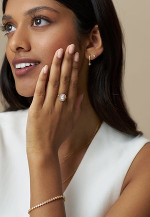 Anello d'oro con una perla ovale circondata da pietre più piccole, indossato su una mano con unghie naturali. Bracciale d'oro sottile e orecchini delicati.