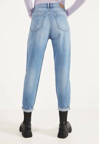 Persona con vaqueros azul claro con puños doblados y ligeras desgastaduras, y botas negras gruesas, posando contra un fondo blanco.