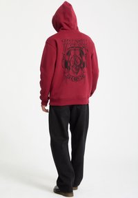 Sudadera burdeos con un diseño gráfico negro que presenta una calavera en la parte posterior, combinada con vaqueros negros y calzado oscuro.