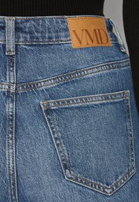 Μπλε τζιν παντελόνι με υφή, που διαθέτει χρυσές ραφές, μια ετικέτα με "VMD" στο πίσω μέρος και κλασικό σχέδιο με πέντε τσέπες.