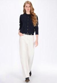 Granatowy sweter z dzianiny z wzorem w warkocze, z okrągłym dekoltem i pięcioma guzikami, w połączeniu z białymi spodniami w szerokim kroju i czarnymi butami.