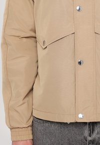 Beige lett jakke laget av glatt materiale, med trykknapper, elastiske mansjetter og frontlommer, supplert med en avslappet passform.