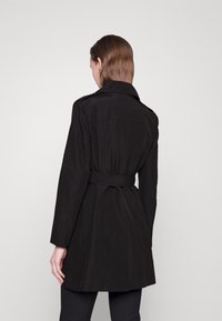 Trench-coat noir avec taille ceinturée, manches longues et coupe cintrée. Tissu lisse, col classique et détail de pli creux à l'arrière.