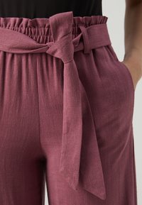 Pantalon large couleur bordeaux en tissu texturé, avec une taille élastique et une ceinture en tissu nouée, conçu avec des poches latérales.