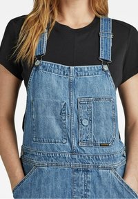 Džínové lacláče s nastavitelnými ramínky, vybavené přední kapsou a dvěma bočními kapsami. Materiál je světle modrý s vybledlým vzhledem.